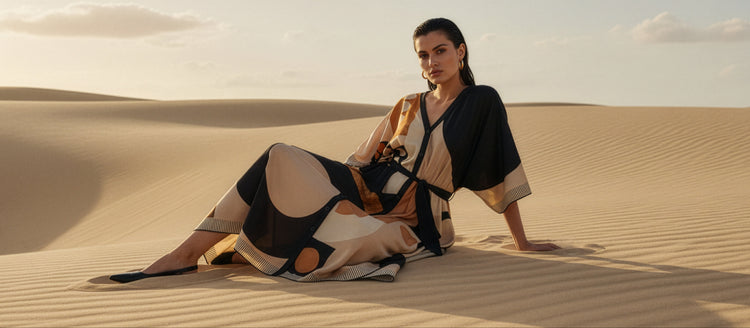 Woman in a stylish graphic colourblock caftandress sitting on sand dunes with a desert landscape.